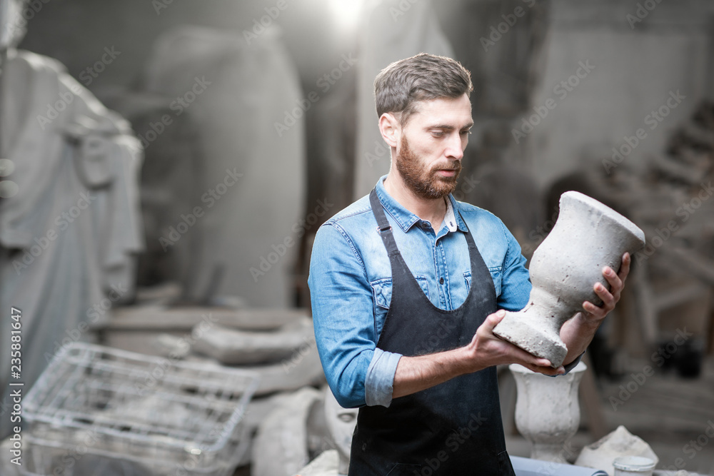 Portrait of a handsome sculptor in blue t-shirt and apron holding old vase in the studio with sculpt