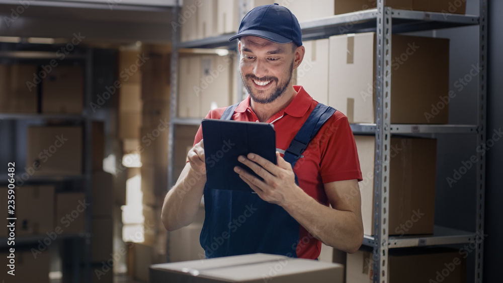 Warehouse Worker Uses Digital Tablet For Checking Stock, On the Shelves Standing Cardboard Boxes.