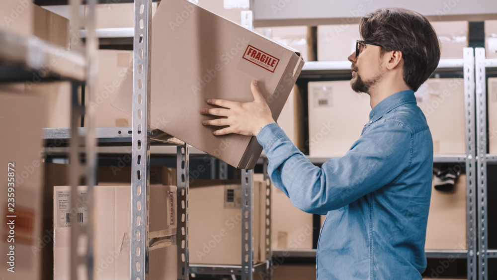 Warehouse Worker Puts Cardboard Box on a Shelf. In the Background Rows of Shelves Full of Cardboard 