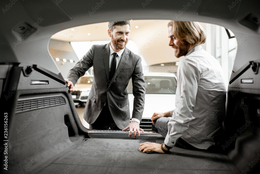 Salesman showing spacious trunk of a new car for a young man client in the showroom