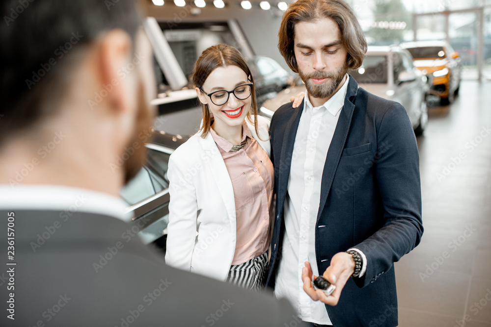 Salesperson giving keys from a new car to a lovely business couple standing together in the car show