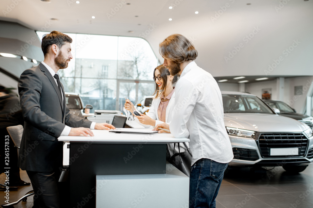 Young business couple with salesperson or manager buying or renting car standing at the stand in the