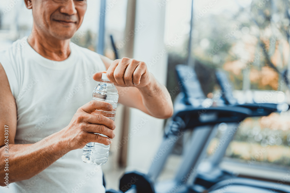 Senior man drink mineral water in gym fitness center after exercise. Elderly healthy lifestyle.