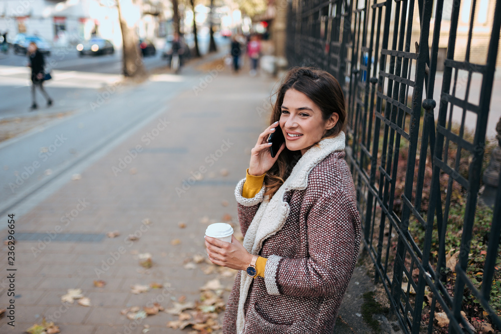 Stylish woman talking on the phone and drinking coffee in the city street.