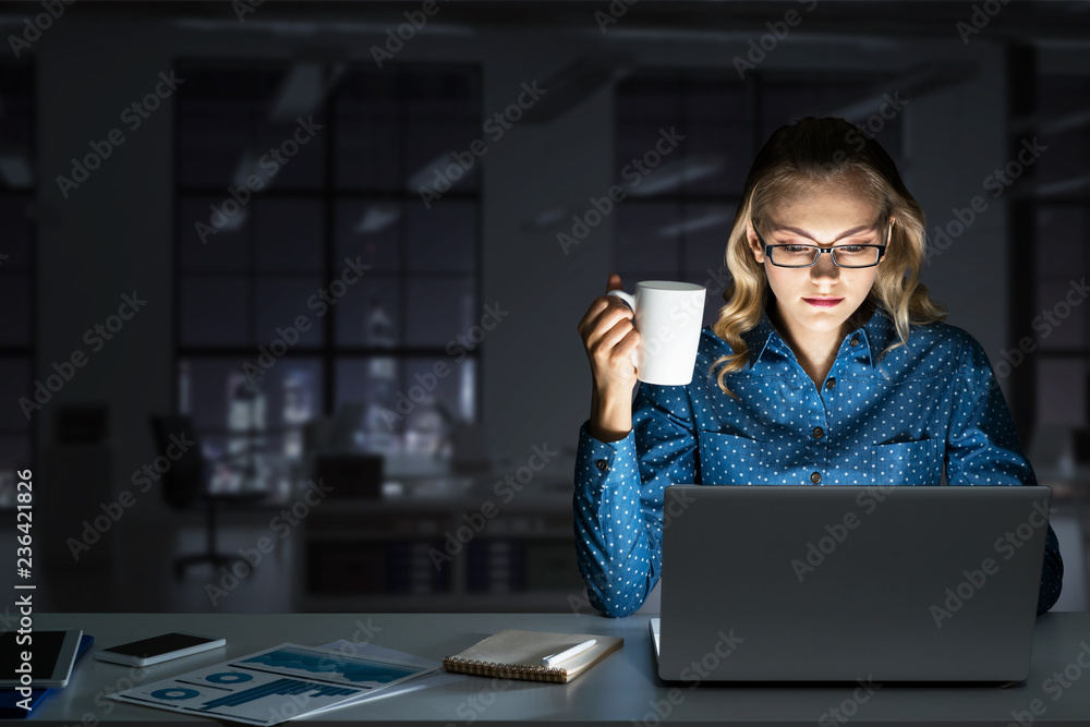 Attractive blonde working on laptop in dark office. Mixed media