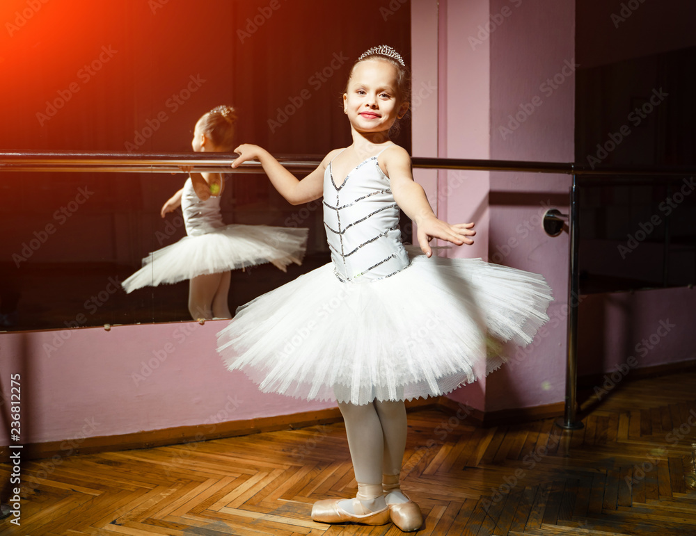 Portrait of smiling young ballerina in white tutu and pointes posing isolated in dance studio. Small