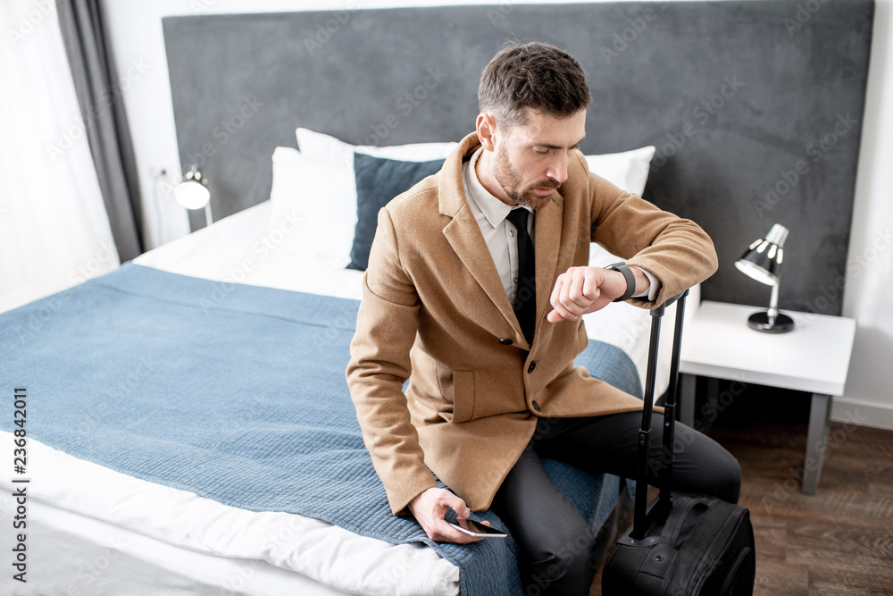 Dressed businessman sitting on the bed with a suitcase watching time before departure from the hotel