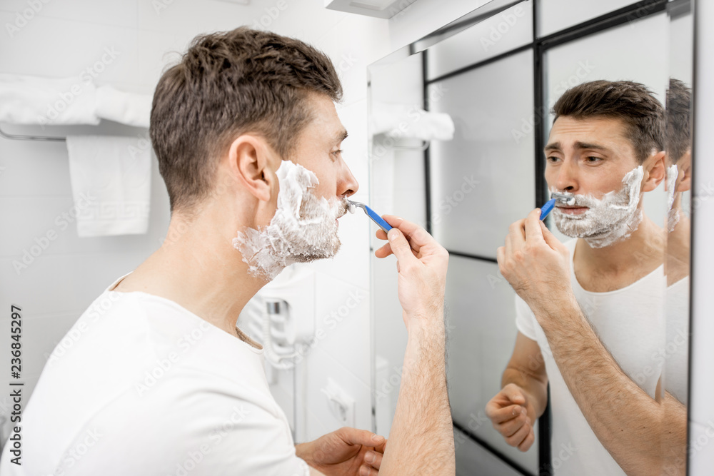 Handsome man in white t-shirt shawing his mustache with blade and foam in the bathroom