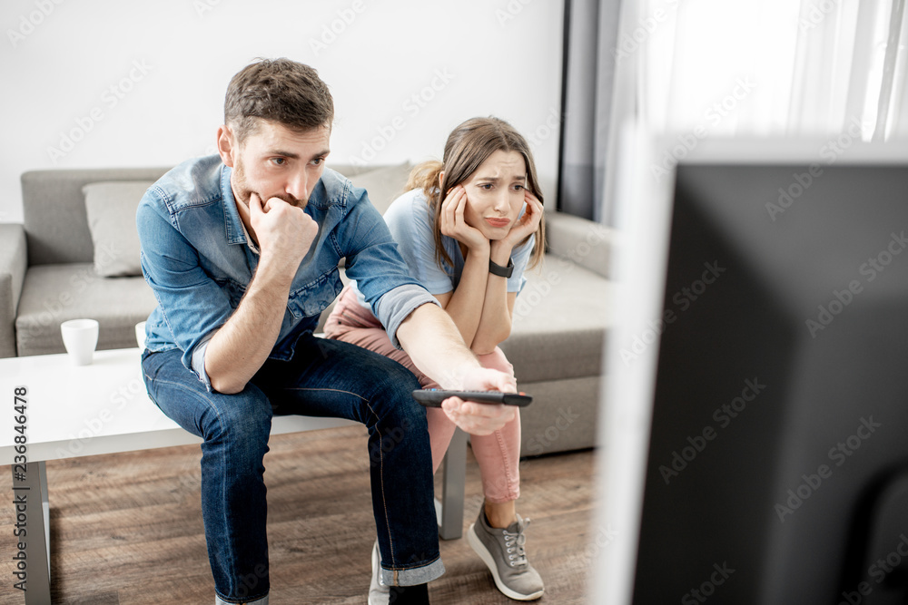 Young couple with unhappy faces watching boring TV sitting together on the table at home