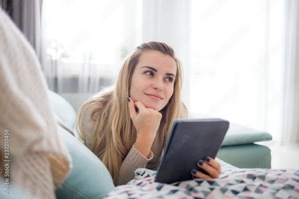 Happy woman reading ebook on digital reader while lying on comfortable sofa at home, leisure time