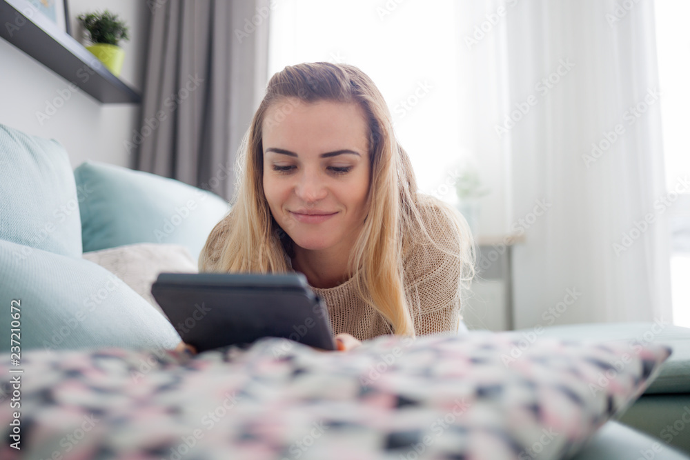 Happy woman reading ebook on digital reader while lying on comfortable sofa at home, leisure time