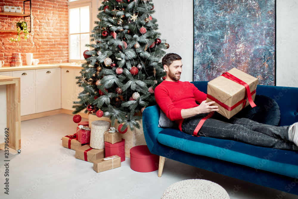 Man opening a gift box lying on the couch during the winter holidays with Christmas tree at home