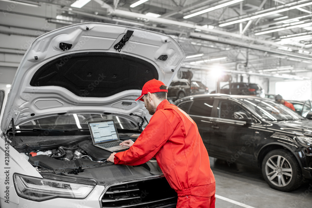 Handsome auto mechanic in red uniform doing engine diagnostics with computer in the car service