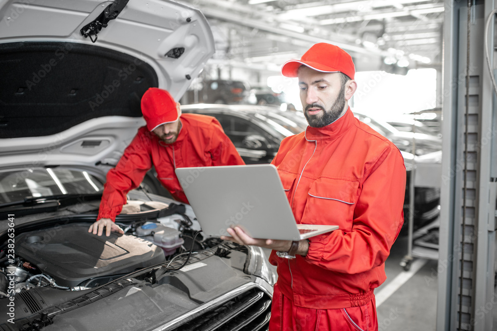 Two auto mechanics in red uniform doing engine diagnostics with computer in the car service