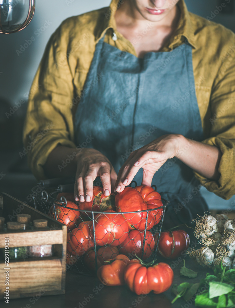 Woman in linen apron taking out from basket ripe heirloom tomatoes for cooking tomato sauce, canned 