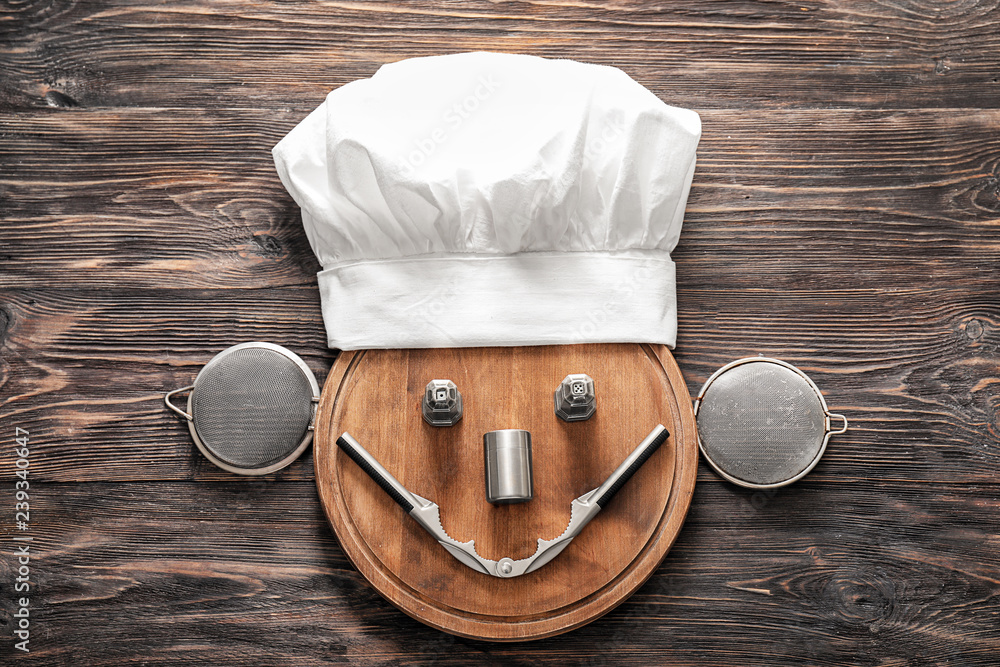 Creative composition with chefs hat, cutting board and kitchen utensils on wooden background