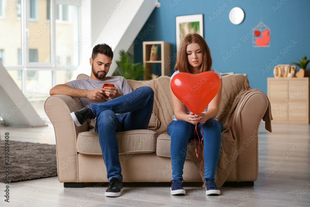 Sad young woman holding heart-shaped balloon near indifferent man playing with mobile phone at home.