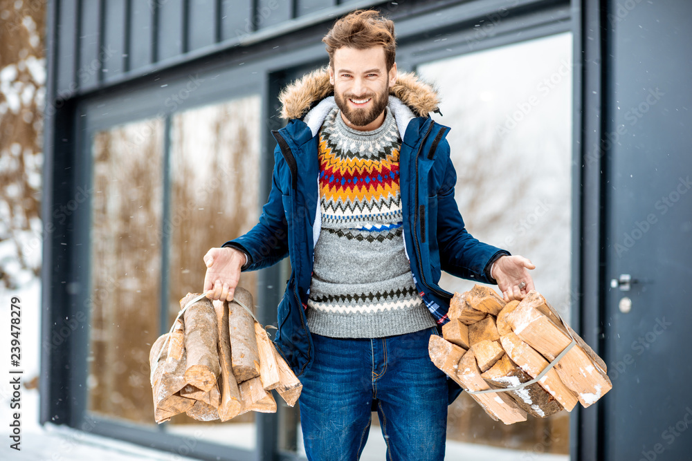 Handsome man in winter clothes carrying firewoods on the terrace near the modern house in the mounta