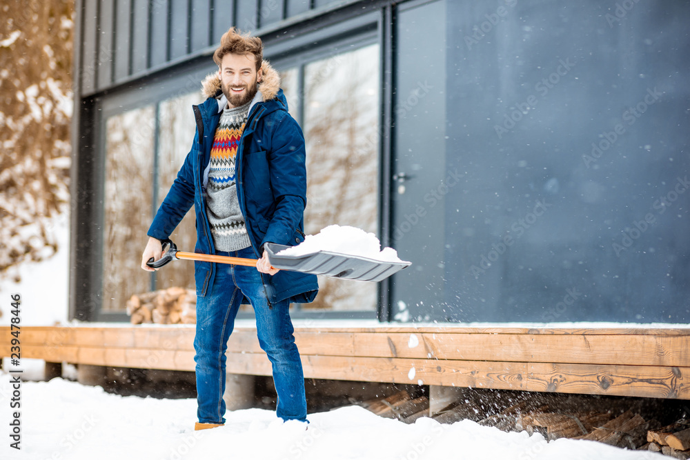 穿着冬装的英俊男子在山里的现代房子附近用铲子清理积雪