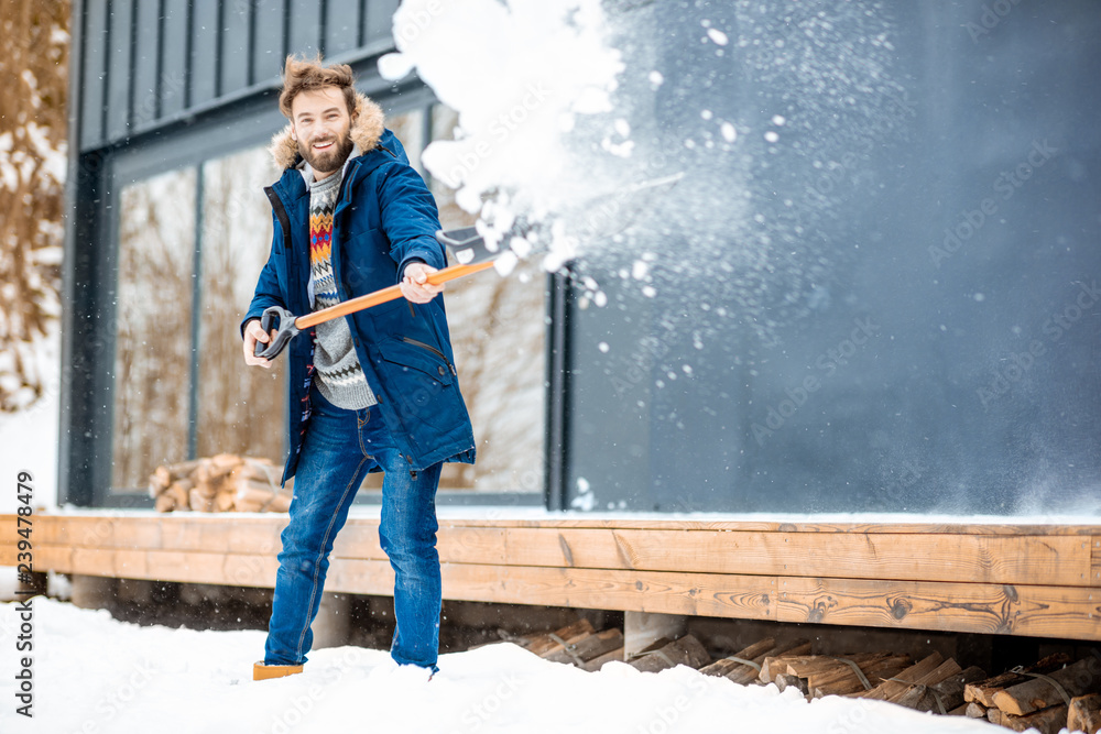 穿着冬装的英俊男子在山里的现代房子附近用铲子清理积雪