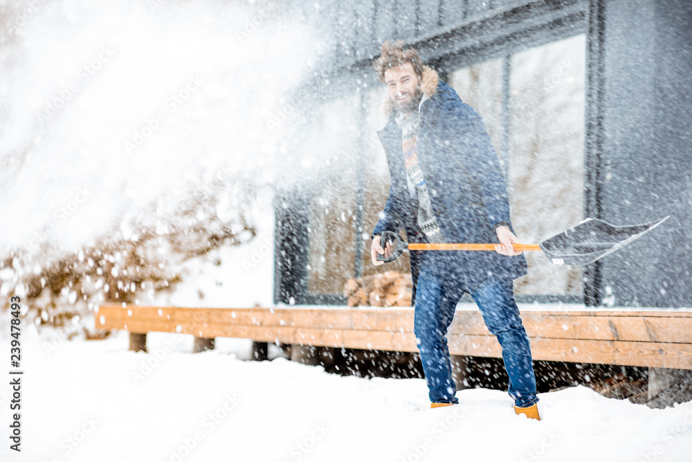 穿着冬装的英俊男子在山里的现代房子附近用铲子清理积雪