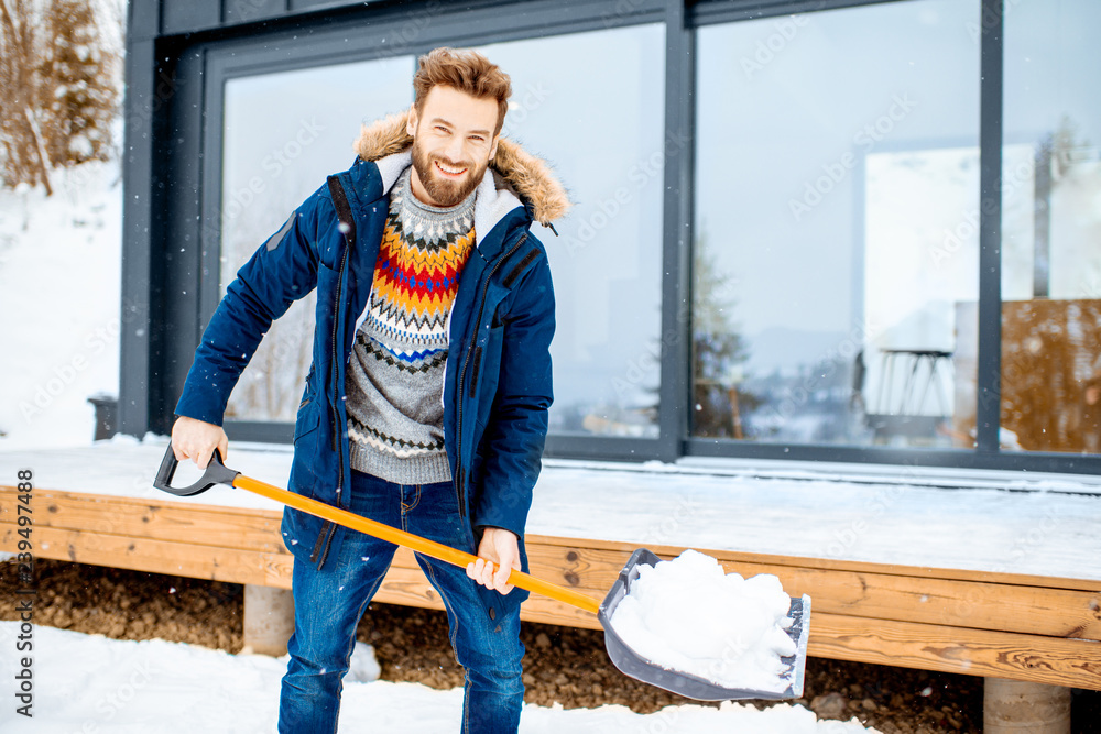 Handsome man in winter clothes cleaning snow with a shovel near the modern house in the mountains