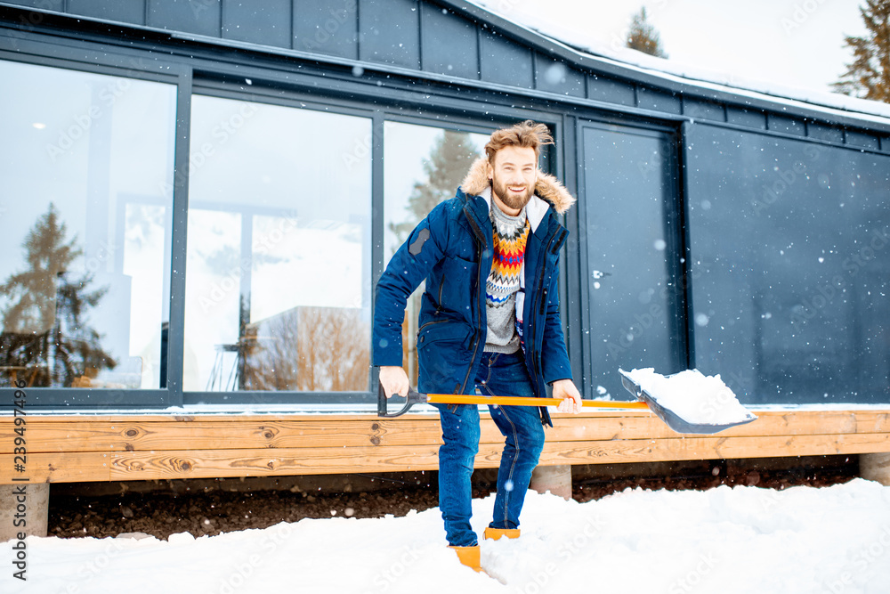 穿着冬装的英俊男子在山里的现代房子附近用铲子清理积雪