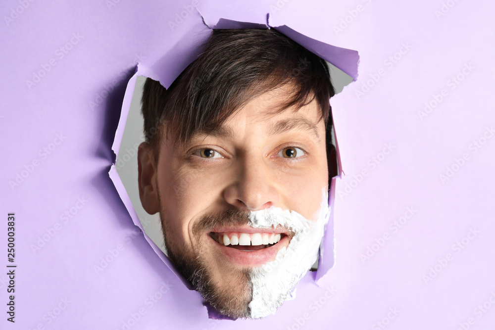 Handsome man with shaving foam on his face visible through hole in torn color paper