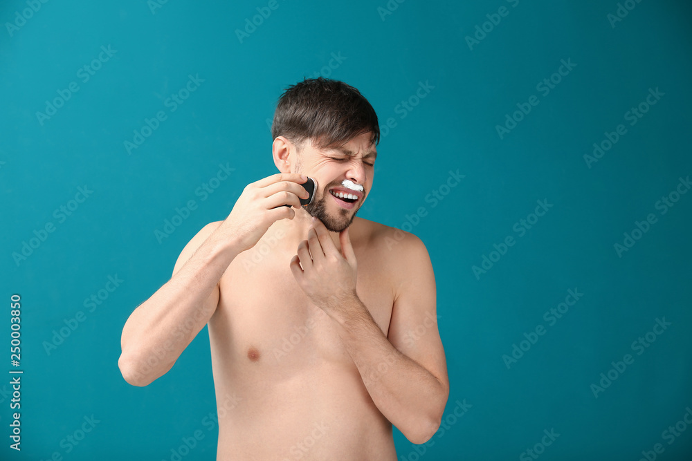 Suffering man using electric shaver against color background