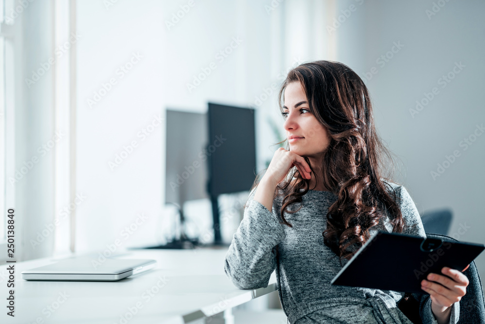 Pensive young woman sitting at workplace holding digital tablet and looking away.