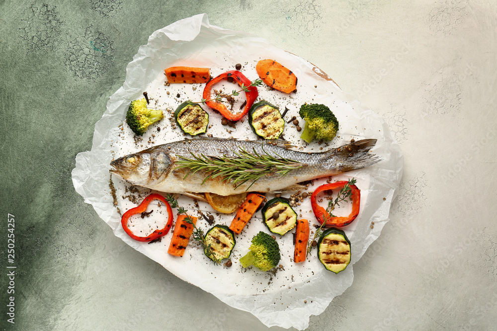 Parchment with tasty cooked seabass fish and vegetables on color background