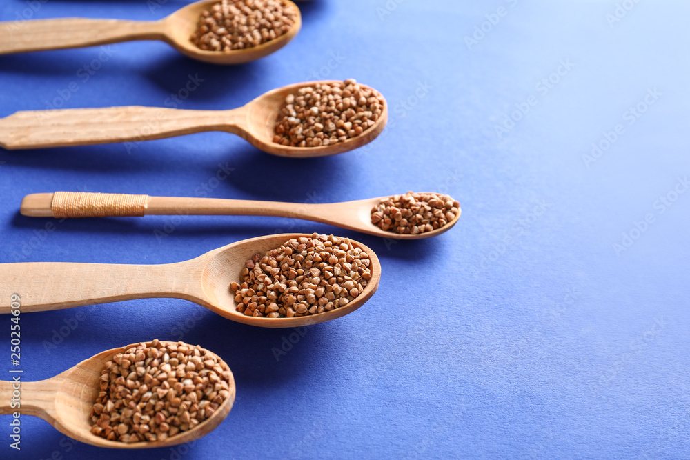 Spoons with raw buckwheat on color background