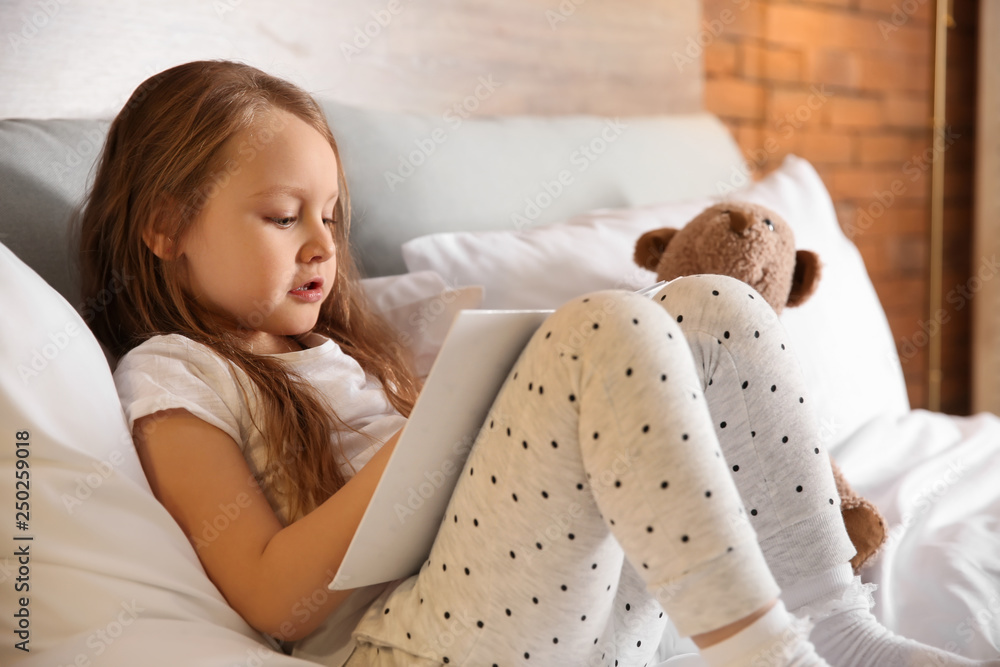 Cute little girl with toy bear reading book in bed