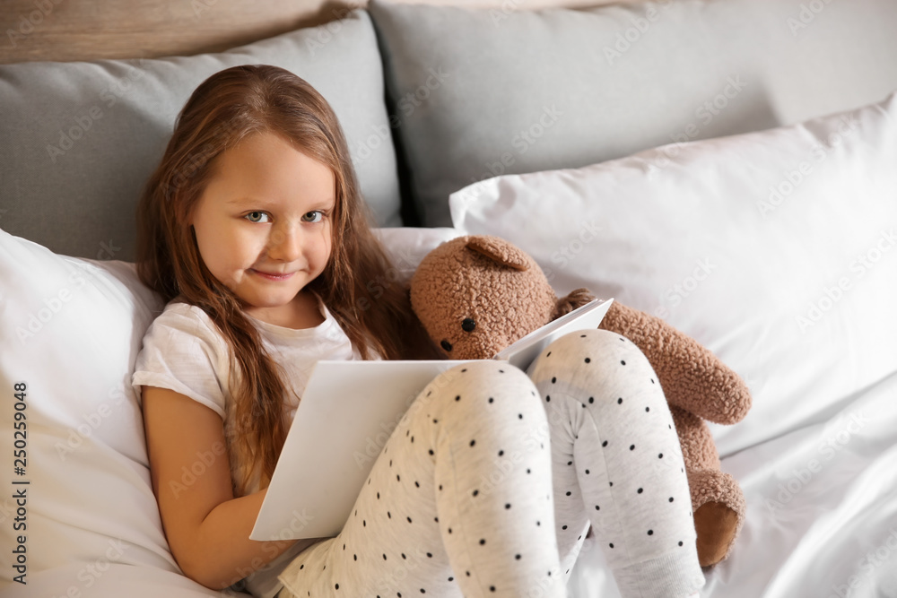 Cute little girl with toy bear reading book in bed