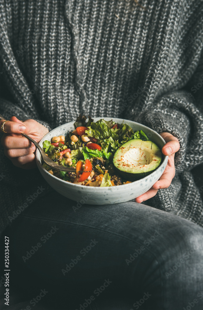 健康的素食晚餐。穿着灰色牛仔裤和保暖毛衣的女人拿着盛有新鲜沙拉和鳄梨的碗