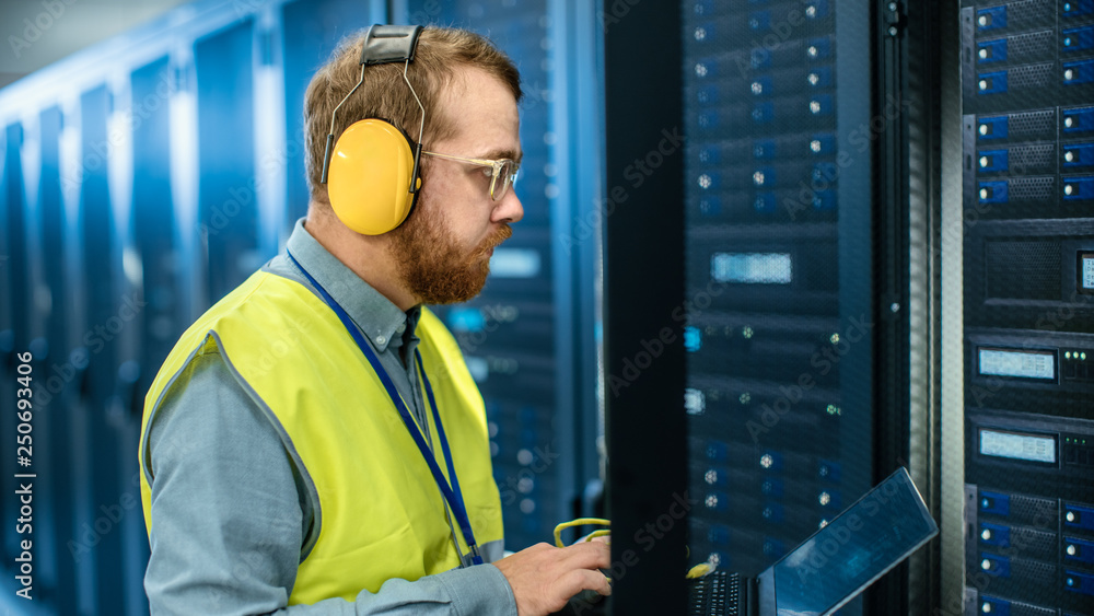 Bearded IT Specialist in Glasses and Headphones, wearing High Visibility Vest is Working on Laptop i