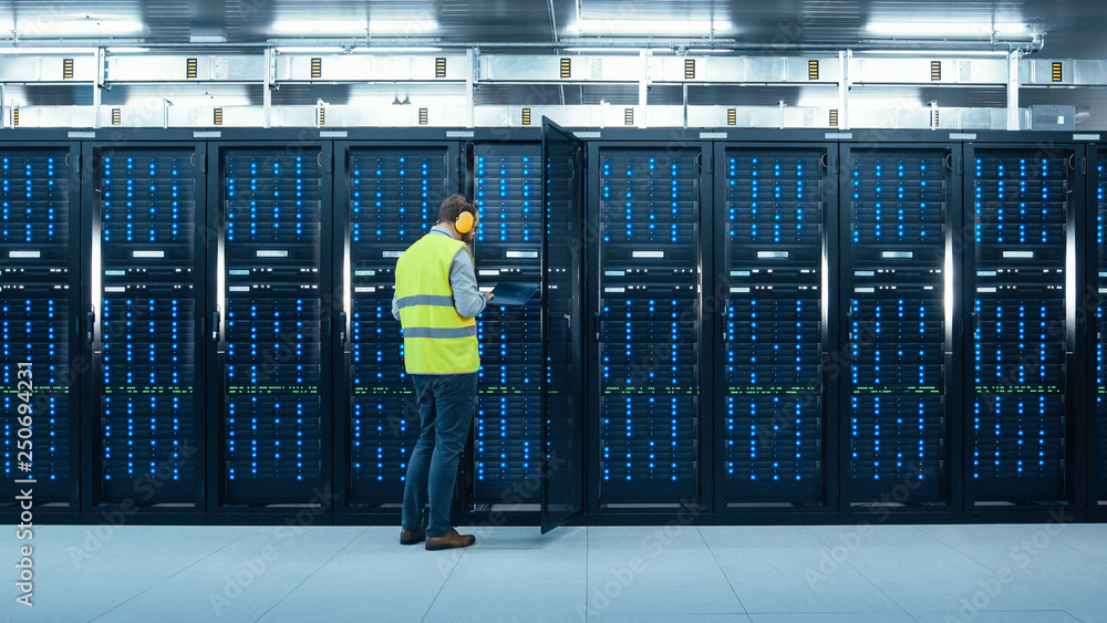 Shot of an IT Engineer in High Visibility Vest and Headphones Working on Laptop in Data Center while