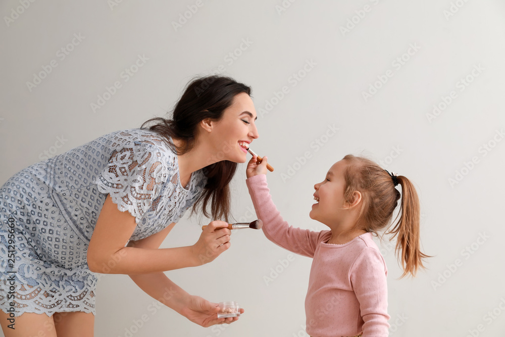 Cute little daughter applying makeup onto mothers face against light background