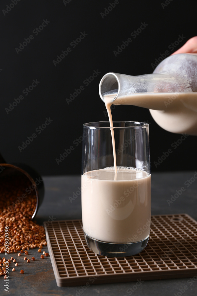 Pouring of tasty buckwheat milk from bottle into glass on dark table