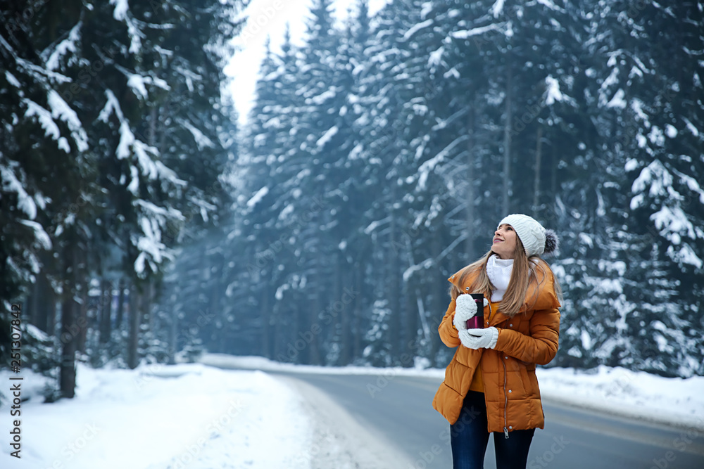 Beautiful young woman with hot drink near road in winter forest