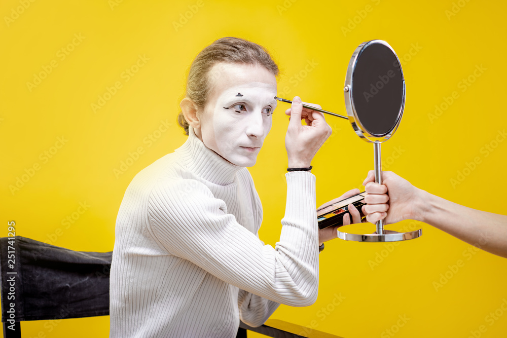 Actor applying facial makeup for pantomime performance sititng in the studio on the yellow backgroun