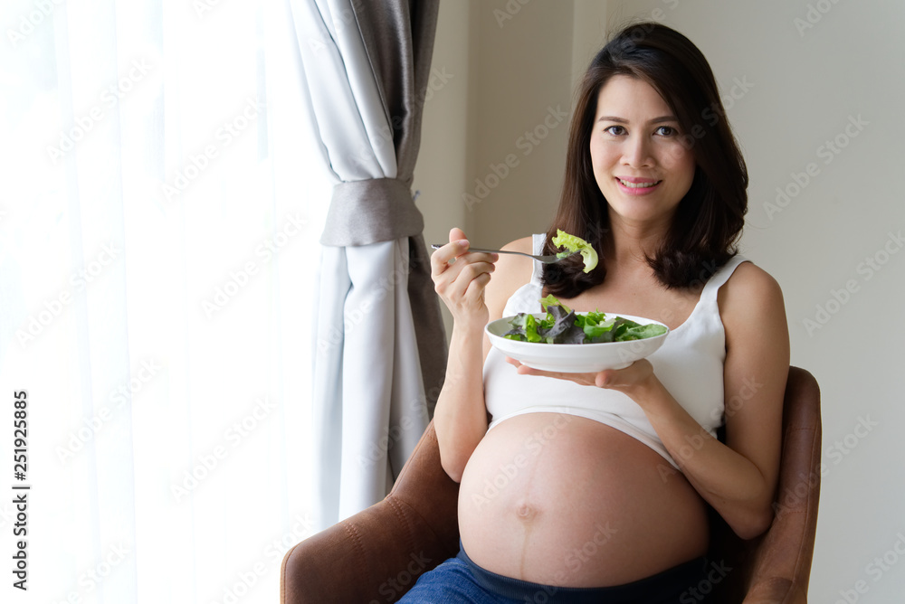 Young Asian pregnant woman eating green salad and vegetables sitting on chair at home. She smiles an