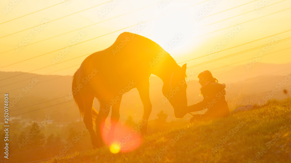 SUN FLARE: Young woman sitting on the hill while her horse grazes at sunrise.