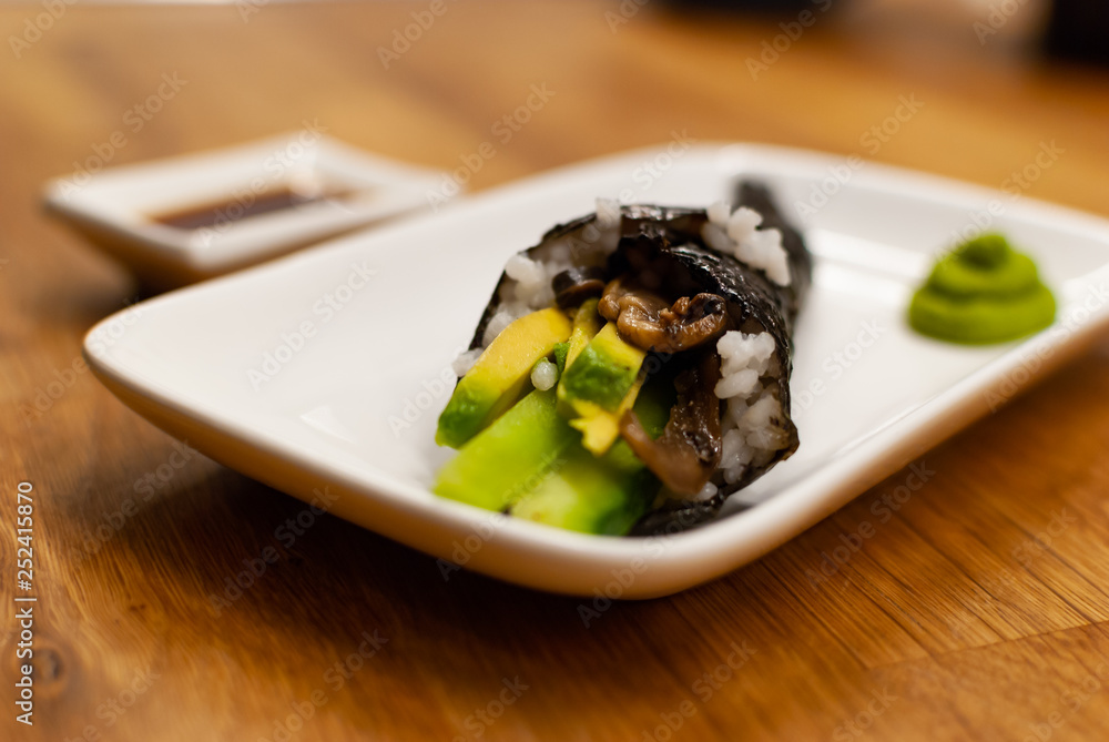 Homemade vegan Temaki-Sushi filled with avocado on a small plate on a wooden table with soy sauce an