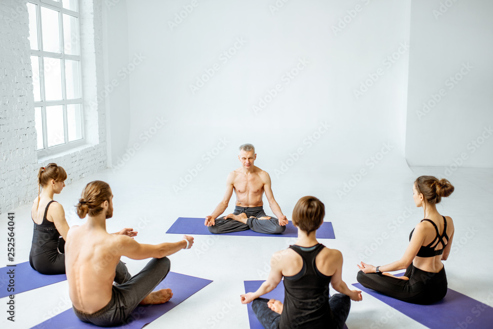 Group of young people practising yoga with experienced senior trainer in the white spacious studio