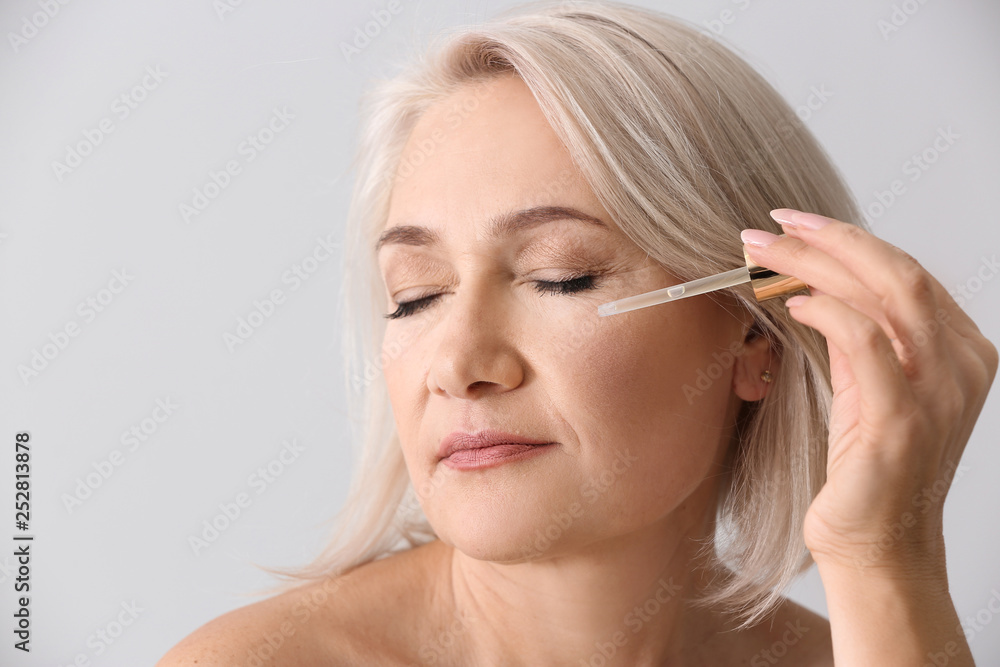 Mature woman applying face serum on light background