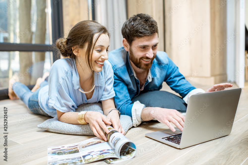 Happy couple lying on the floor with laptop and magazines, relaxing at the cozy home