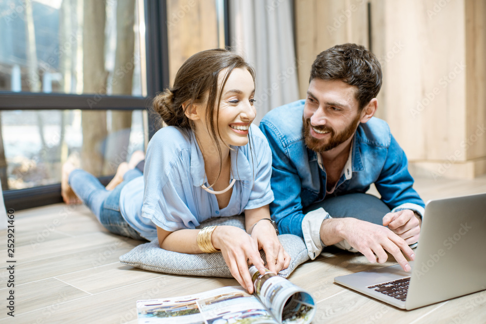 Happy couple lying on the floor with laptop and magazines, relaxing at the cozy home