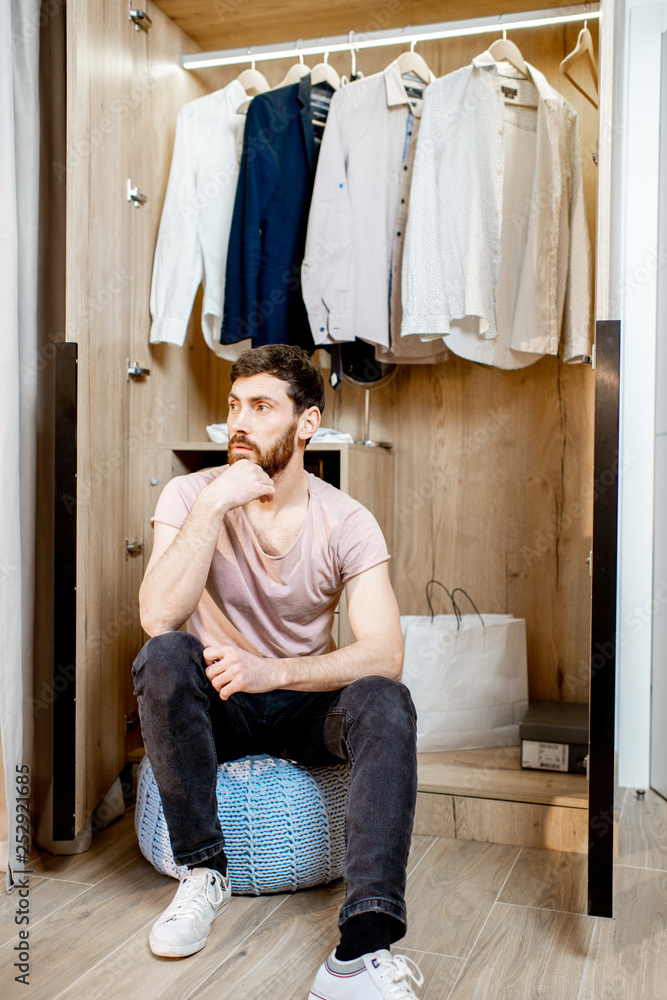 Handsome man sitting in the wardrobe full of clothes at home