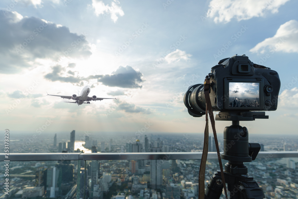 Black full frame camera on tripod take photograph of airplane take off with city view